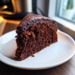 A close-up of a moist slice of Gluten-Free Chocolate Cake with dark chocolate frosting on a white plate.