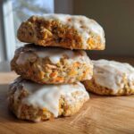 A stack of three Soft-Baked Carrot Cake Cookies with Cream Cheese Glaze, with one cookie resting beside them on a wooden surface.