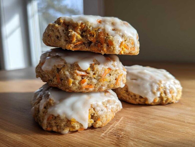 A stack of three Soft-Baked Carrot Cake Cookies with Cream Cheese Glaze, with one cookie resting beside them on a wooden surface.