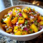 Close-up of a white bowl filled with bright yellow, diced mango mixed with purple red onion and green cilantro, showcasing the Spicy Mango Salsa.