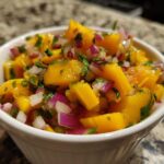 Close-up of vibrant Spicy Mango Salsa featuring chunks of yellow mango, red onion, and cilantro in a white bowl.