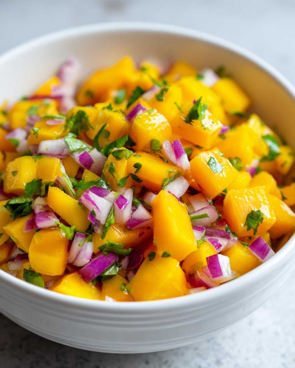 Close-up of bright yellow cubes of mango mixed with chopped red onion and cilantro in a white bowl, showcasing the Spicy Mango Salsa.