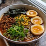 A bowl of Spicy Miso Ramen featuring rich broth, ground meat, corn, scallions, nori, and three halves of soft-boiled eggs.