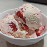 Close-up of creamy Strawberry Banana Ice Cream scoops swirled with fresh strawberry pieces in a white bowl.