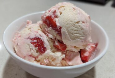 Close-up of creamy Strawberry Banana Ice Cream scoops swirled with fresh strawberry pieces in a white bowl.