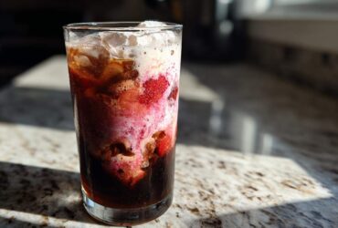A tall glass of layered Strawberry Cream Cold Brew with visible strawberries and foam, sitting on a granite counter in bright sunlight.