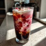 Close-up of a tall glass of Strawberry Cream Cold Brew showing dark coffee, pink/red strawberry syrup, and creamy white foam topping.