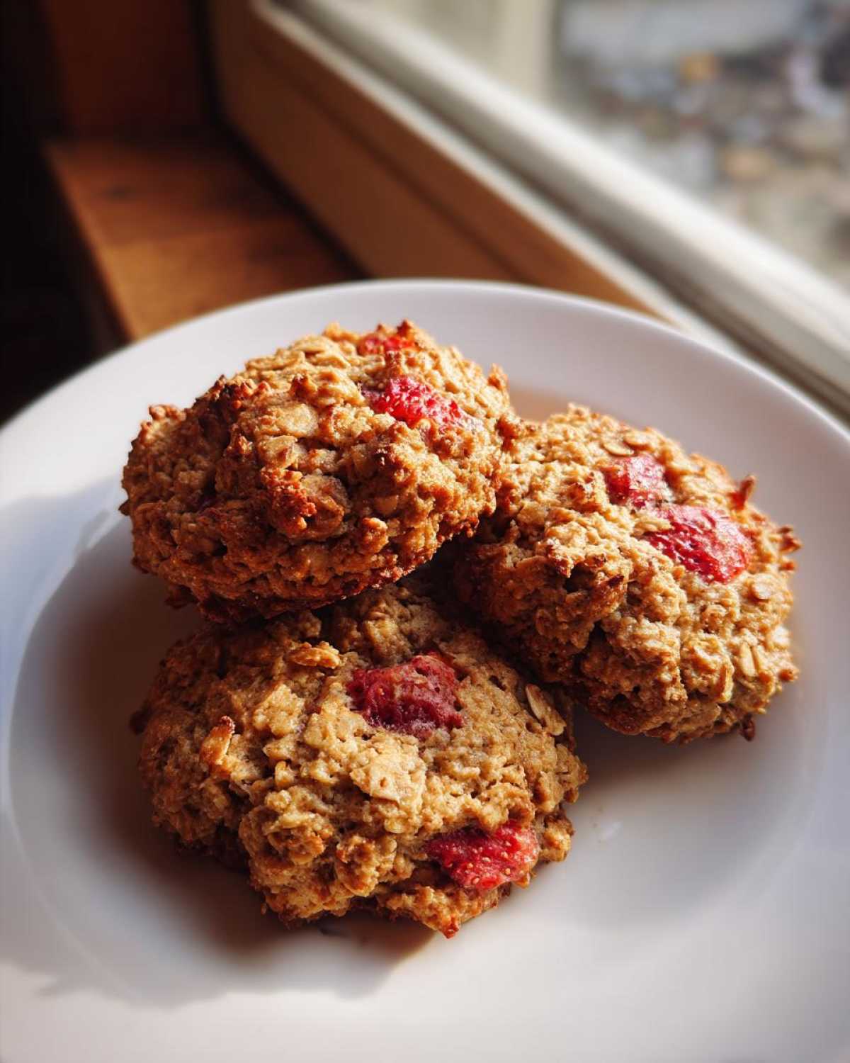 Three golden brown Strawberry Oatmeal Breakfast Cookies stacked slightly on a white plate near a sunny window.
