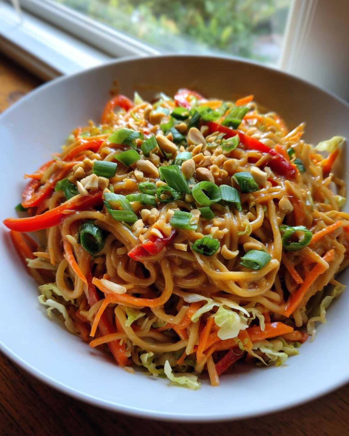 Close-up of a white bowl filled with saucy Thai Peanut Noodles with Crunchy Veggies, topped with peanuts and green onions.