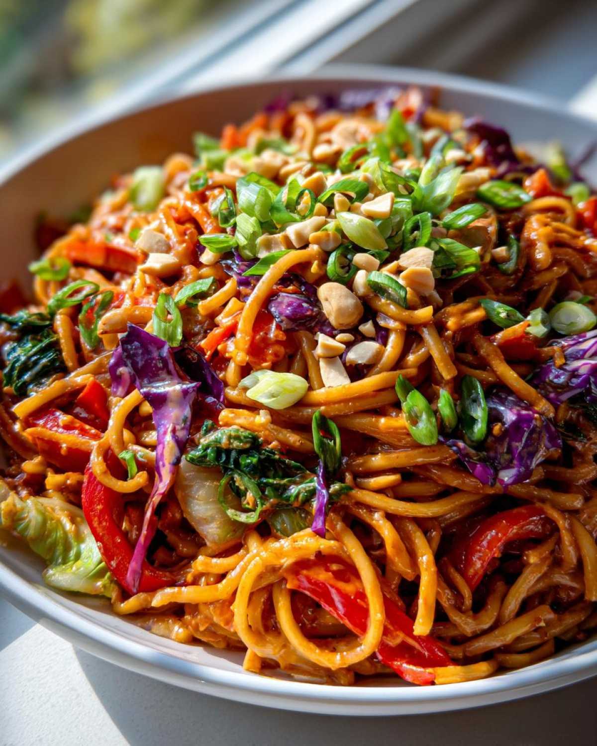 Close-up of a bowl of rich Thai Peanut Noodles with Crunchy Veggies, topped with peanuts and green onions.