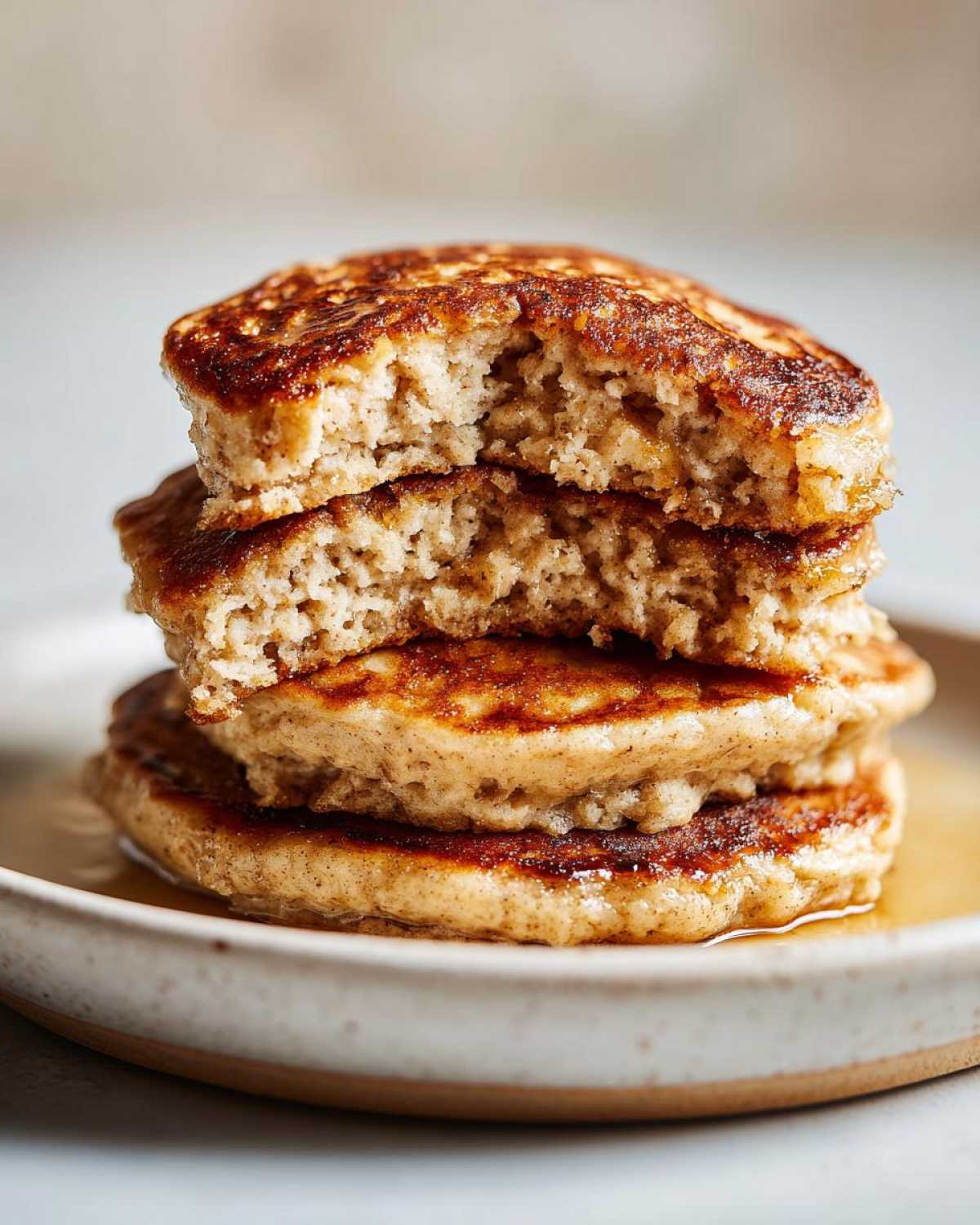 Close-up of a stack of fluffy Vegan Banana Oat Pancakes, one bitten into, drizzled with syrup.