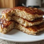 A stack of three thick Vegan Banana Oat Pancakes, cut open to show the texture of oats inside, served on a white plate.