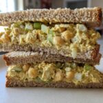 A cross-section view of a delicious Vegan Chickpea Salad Sandwich stacked on a white plate, showing creamy filling and celery chunks.