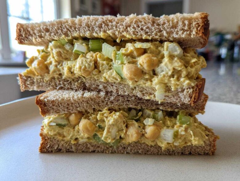 A cross-section view of a delicious Vegan Chickpea Salad Sandwich stacked on a white plate, showing creamy filling and celery chunks.