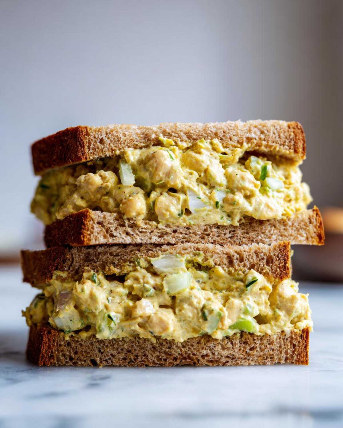 Two halves of a Vegan Chickpea Salad Sandwich stacked, featuring creamy yellow chickpea filling on dark whole wheat bread.