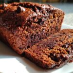 Close-up of a rich, dark Vegan Chocolate Banana Bread loaf with one slice cut, showing moist texture and chocolate chips.