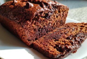 Close-up of a rich, dark Vegan Chocolate Banana Bread loaf with one slice cut, showing moist texture and chocolate chips.