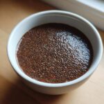 Overhead close-up of thick Vegan Chocolate Chia Pudding in a small white bowl on a wooden surface.