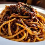 Close-up of thick spaghetti noodles coated in a rich, dark Vegan Lentil Bolognese sauce, garnished with herbs.