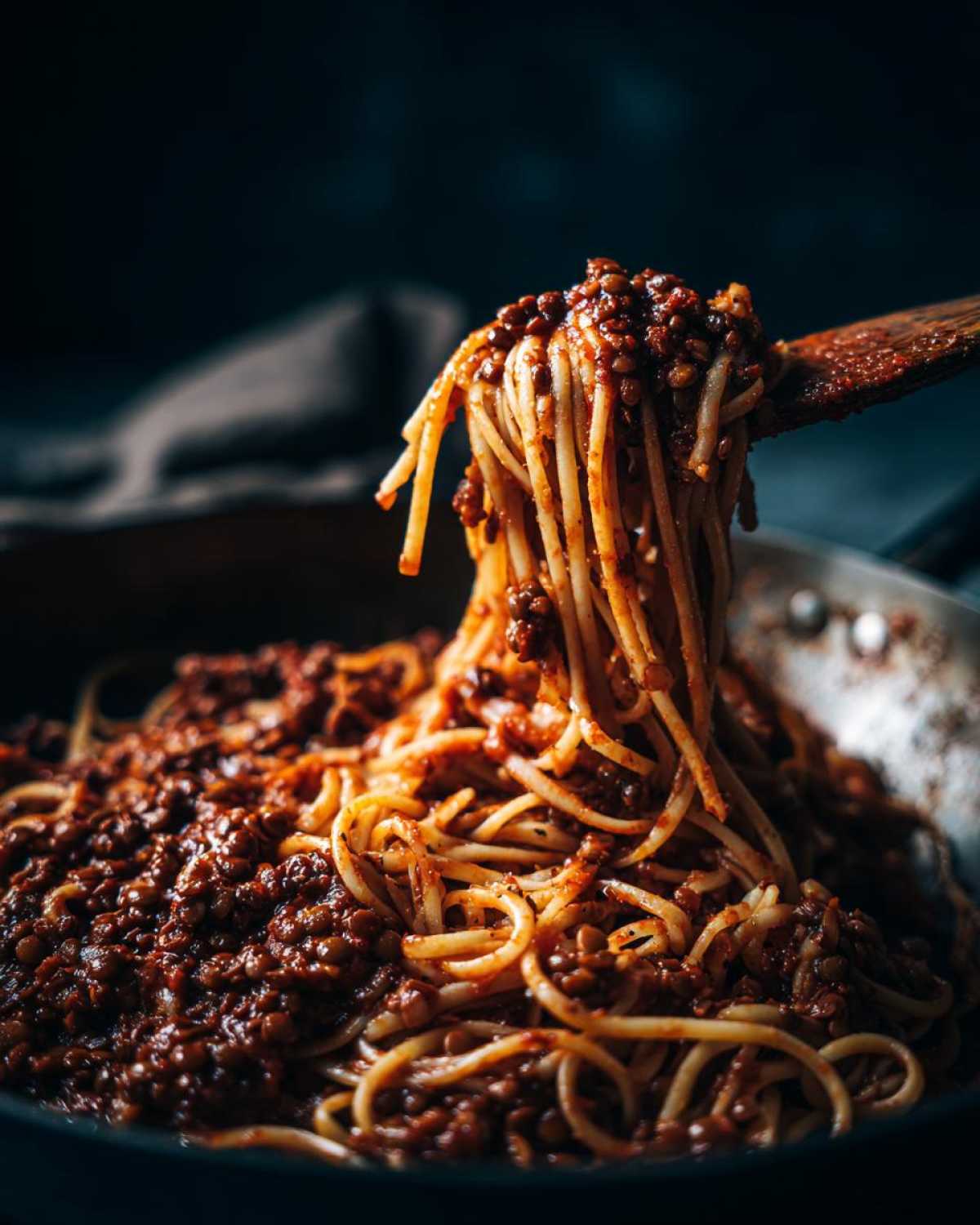 A wooden spoon lifts spaghetti coated in rich Vegan Lentil Bolognese sauce from a pan.