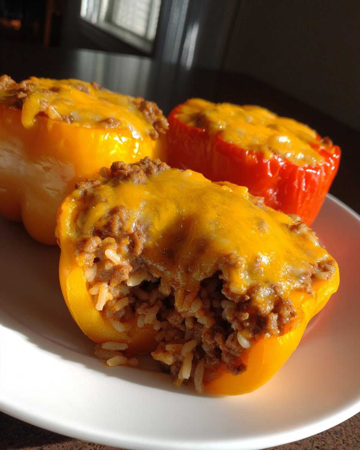 Three baked stuffed bell peppers, two yellow and one red, topped with melted cheddar cheese, showing the ground beef and rice filling.