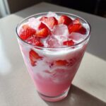 Close-up of a glass filled with a pink beverage, ice, and sliced strawberries, resembling a Copycat Starbucks Pink Drink.