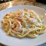Close-up of a white plate holding a generous serving of Creamy Alfredo Shrimp Pasta with Garlic, topped with parmesan and pepper.