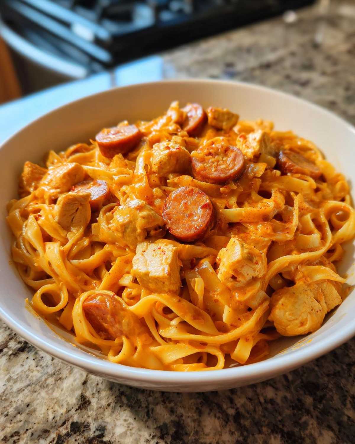 A close-up of a white bowl filled with Creamy Cajun Chicken Pasta featuring fettuccine, chicken chunks, and sliced andouille sausage.