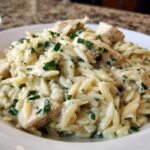 A close-up of a bowl of Creamy Lemon Chicken Orzo mixed with chunks of chicken and topped with fresh green herbs.