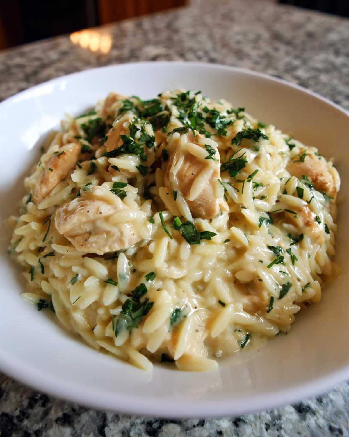 A close-up of a white bowl filled with creamy Lemon Chicken Orzo, topped generously with fresh chopped parsley.