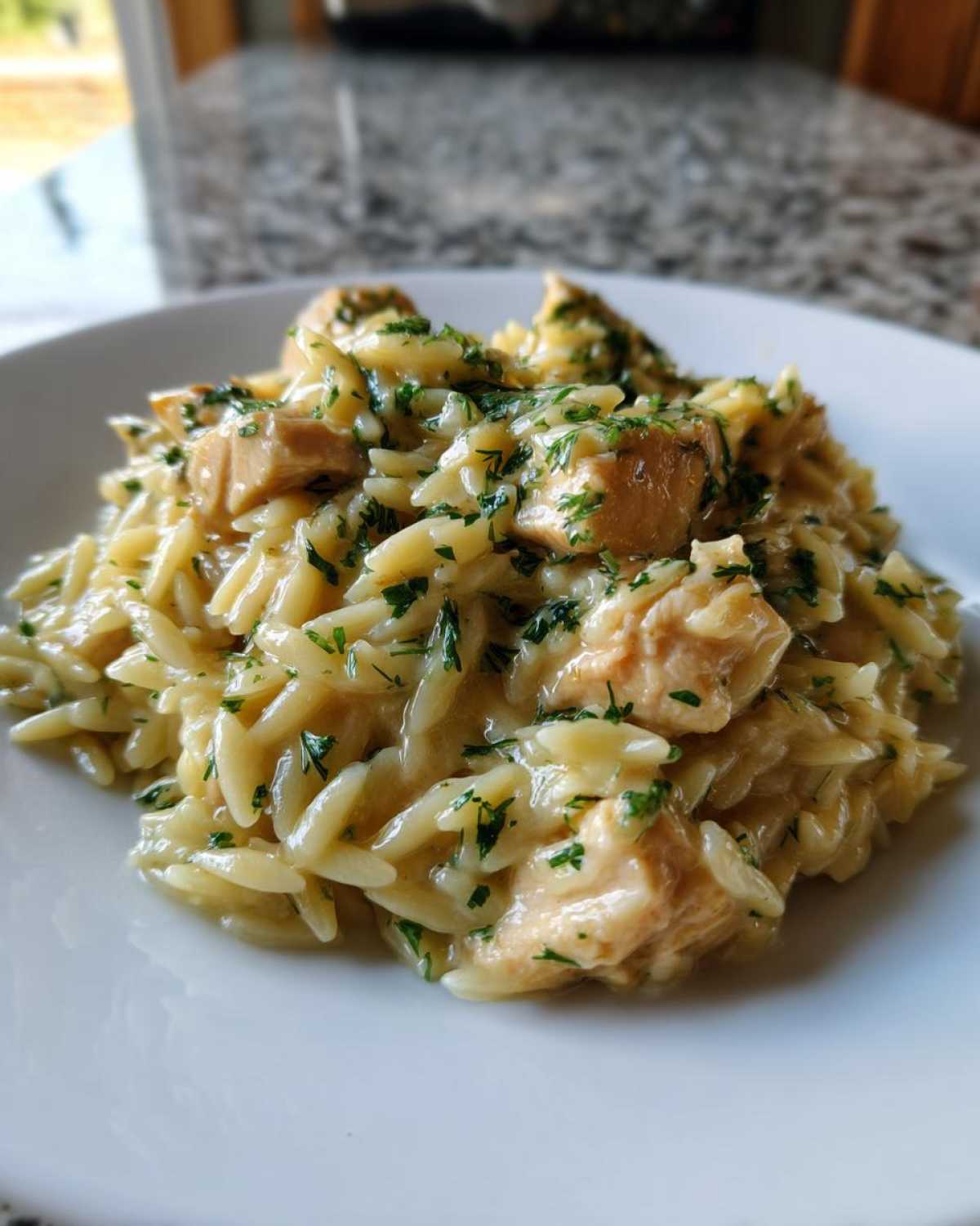 A close-up of a serving of Creamy Lemon Chicken Orzo with visible chunks of chicken and fresh green herbs.
