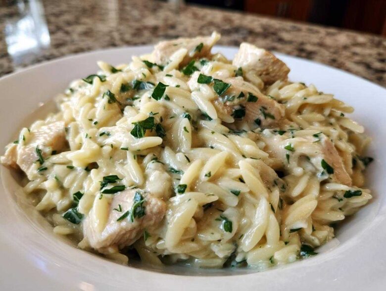 A close-up of a bowl of Creamy Lemon Chicken Orzo mixed with chunks of chicken and topped with fresh green herbs.