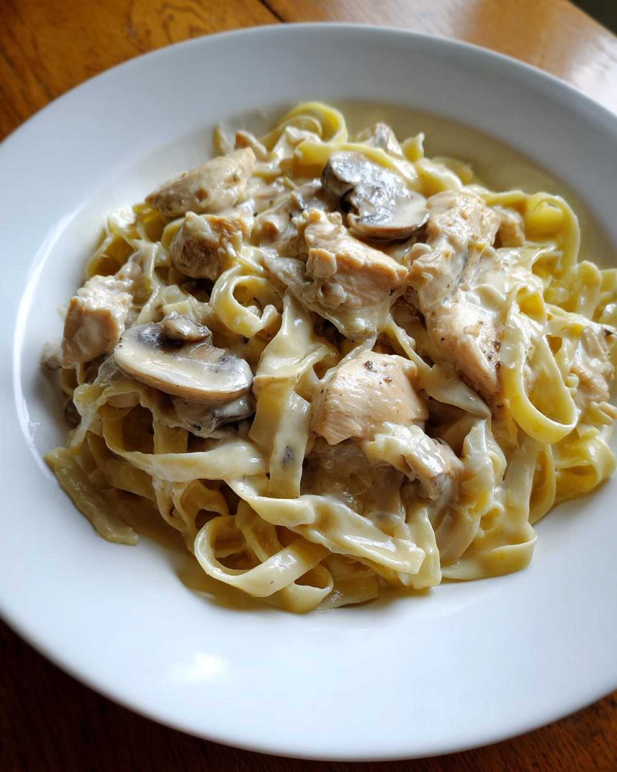 A close-up of a white bowl filled with rich Creamy Mushroom Chicken Alfredo Pasta featuring fettuccine noodles.