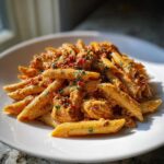 A close-up of a serving of Creamy Sun Dried Tomato Pasta with Chicken, coated in a rich orange sauce and garnished with parsley.