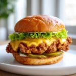 A close-up of a towering Crispy Chicken Burger with melted cheese, lettuce, and pickles on a brioche bun.