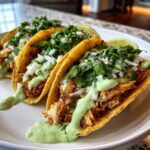 Close-up of three Crispy Chicken Tacos topped with avocado cream, cilantro, and onions, served on a white plate.