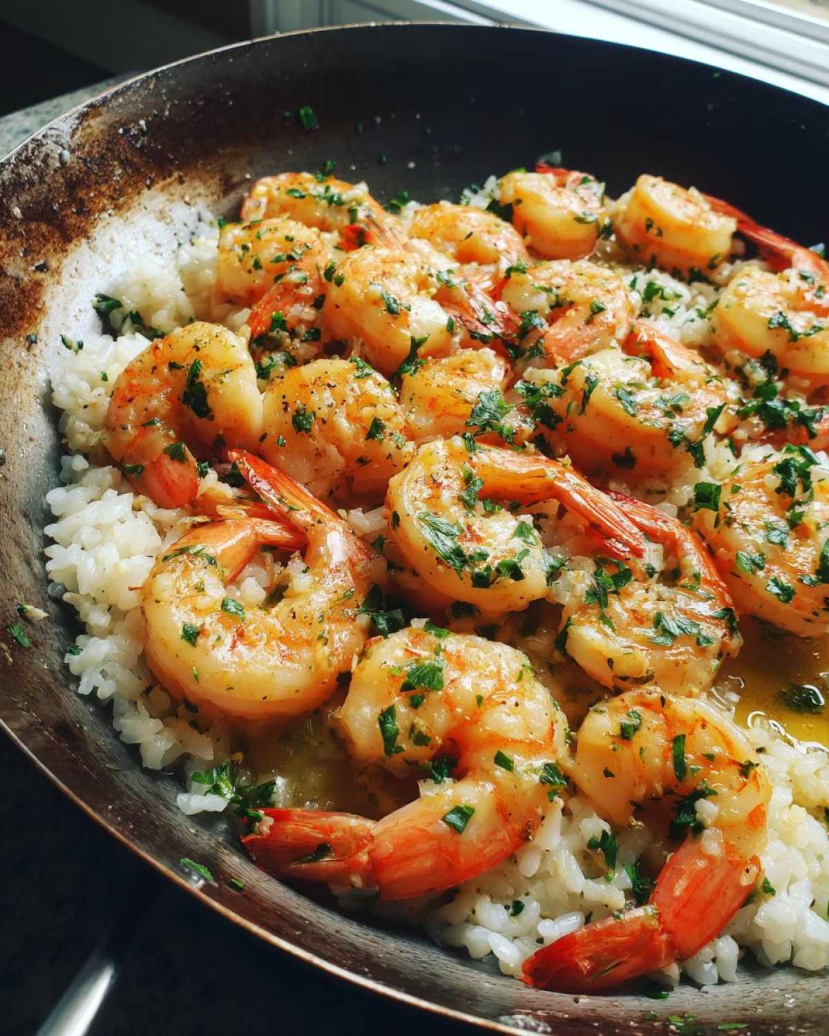 Close-up of plump, cooked shrimp coated in garlic butter sauce and parsley served over white rice in a dark skillet.