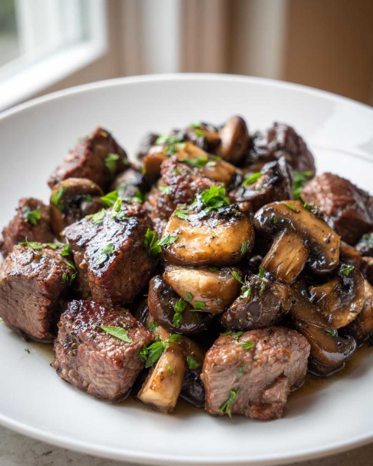 Close-up of savory Garlic Butter Steak Bites with Mushrooms, garnished with fresh parsley, served on a white plate.