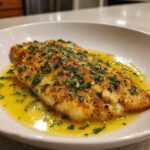 A beautifully pan-seared fillet of Garlic Butter Tilapia swimming in a rich, bright yellow lemon butter sauce, topped with fresh parsley.