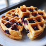 Close-up of golden Gluten-Free Blueberry Waffles, one broken open, showing juicy blueberries baked inside.