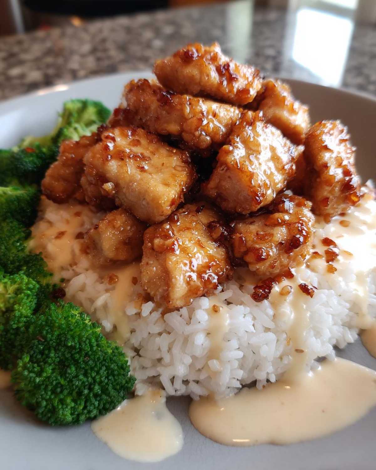 Close-up of a Honey Garlic Chicken Bowl featuring glazed chicken pieces over white rice, drizzled with creamy sauce, and served with steamed broccoli.
