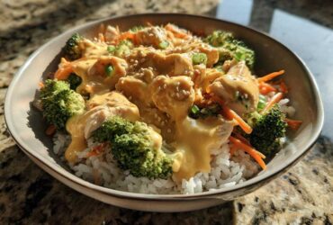 A close-up of a Honey Mustard Chicken Bowl featuring chicken pieces, broccoli, carrots, and rice topped with a creamy sauce and sesame seeds.