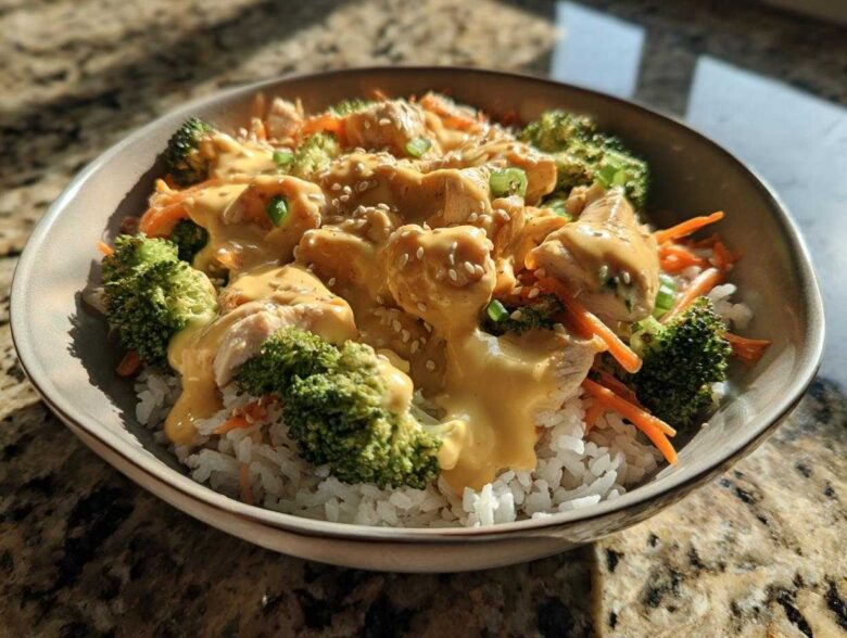 A close-up of a Honey Mustard Chicken Bowl featuring chicken pieces, broccoli, carrots, and rice topped with a creamy sauce and sesame seeds.