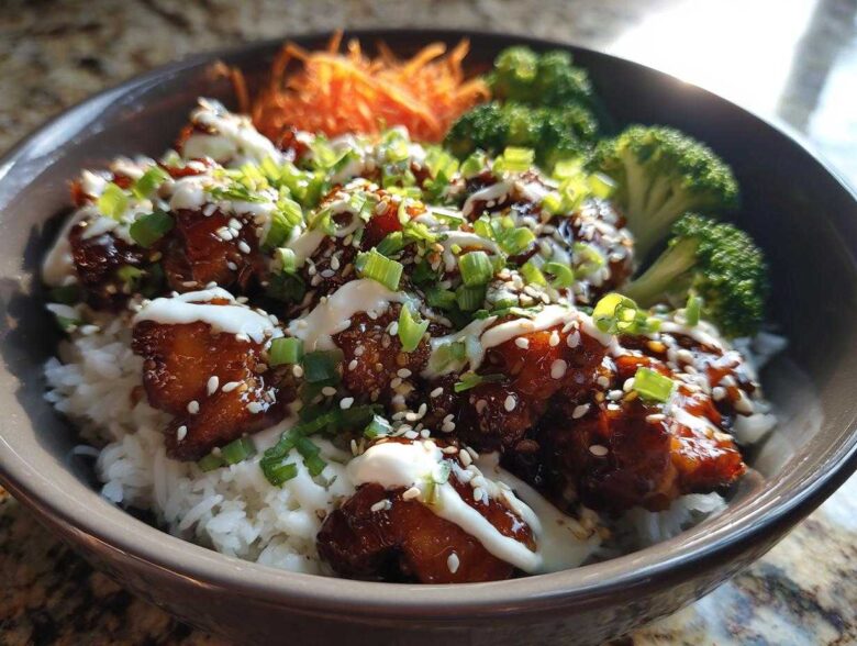 Close-up of a Honey Soy Chicken Bowl featuring glazed chicken pieces over white rice, topped with white sauce, sesame seeds, and green onions.