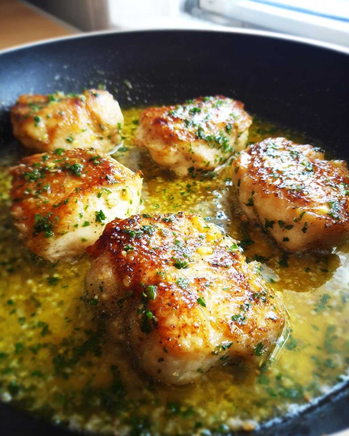 Four pieces of golden-brown chicken simmering in a rich lemon garlic butter sauce with parsley in a black skillet.