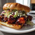 Close-up of a Sticky BBQ Chicken Sandwich with chunks of glazed chicken and crunchy slaw on a brioche bun.