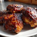 Close-up of four glistening Sticky BBQ Chicken Thighs with crispy, caramelized edges served on a white plate.