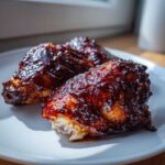 Three pieces of juicy chicken thighs covered in a thick, dark, sticky BBQ chicken thighs with honey glaze, served on a white plate.