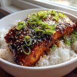 A close-up of a Sticky Honey Soy Salmon with Rice Bowl, featuring glazed salmon topped with sesame seeds and green onions.