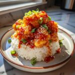 A mound of white rice topped with glazed, crispy chicken coated in sticky sweet chili sauce, garnished with sesame seeds and scallions.